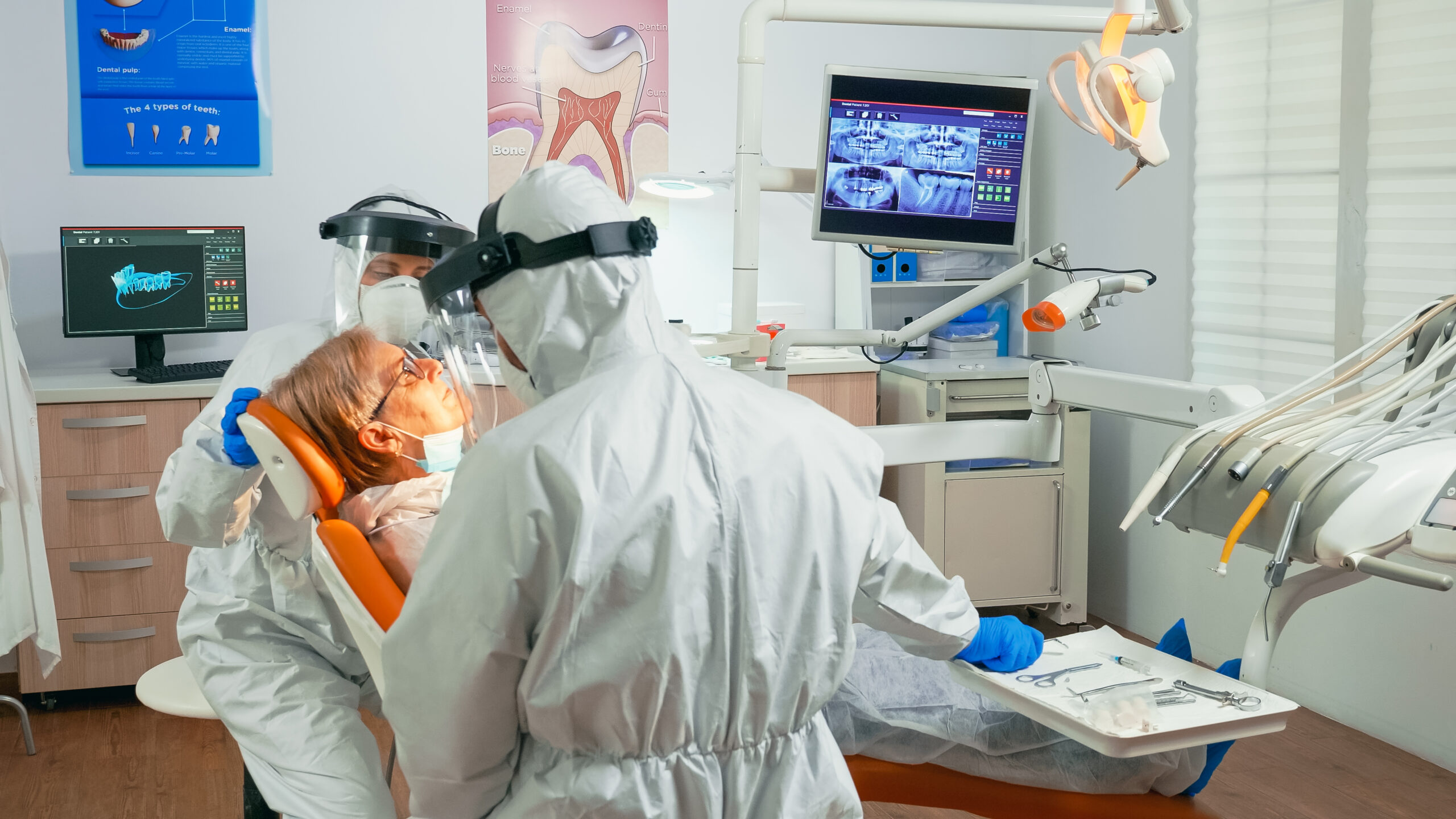 Uma enfermeira e um médico, ambos usando trajes de proteção, estavam trabalhando na unidade odontológica durante a pandemia de coronavírus. Eles participavam do tratamento de pacientes, incluindo um assistente de paciente sênior e um médico ortodôntico. Todos usavam macacões, protetores faciais, máscaras e luvas como parte das medidas de segurança.