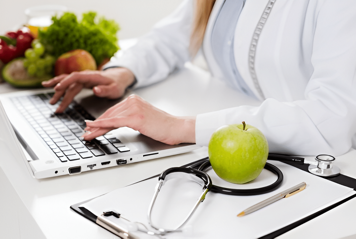 Nutricionista digitando no notebook ao lado de frutas e legumes.